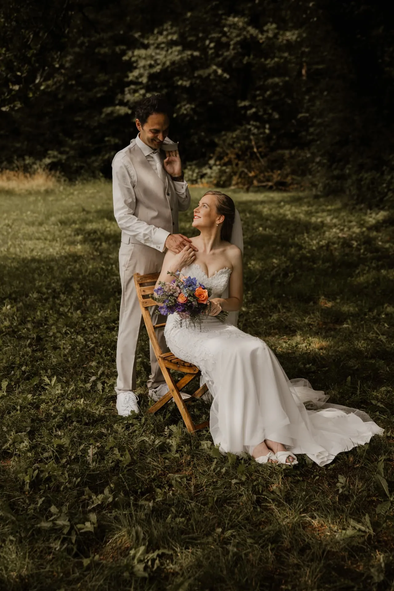 Bruid op een houten stoel in het gras met bruidegom achter haar, trouwfotografie Achterhoek