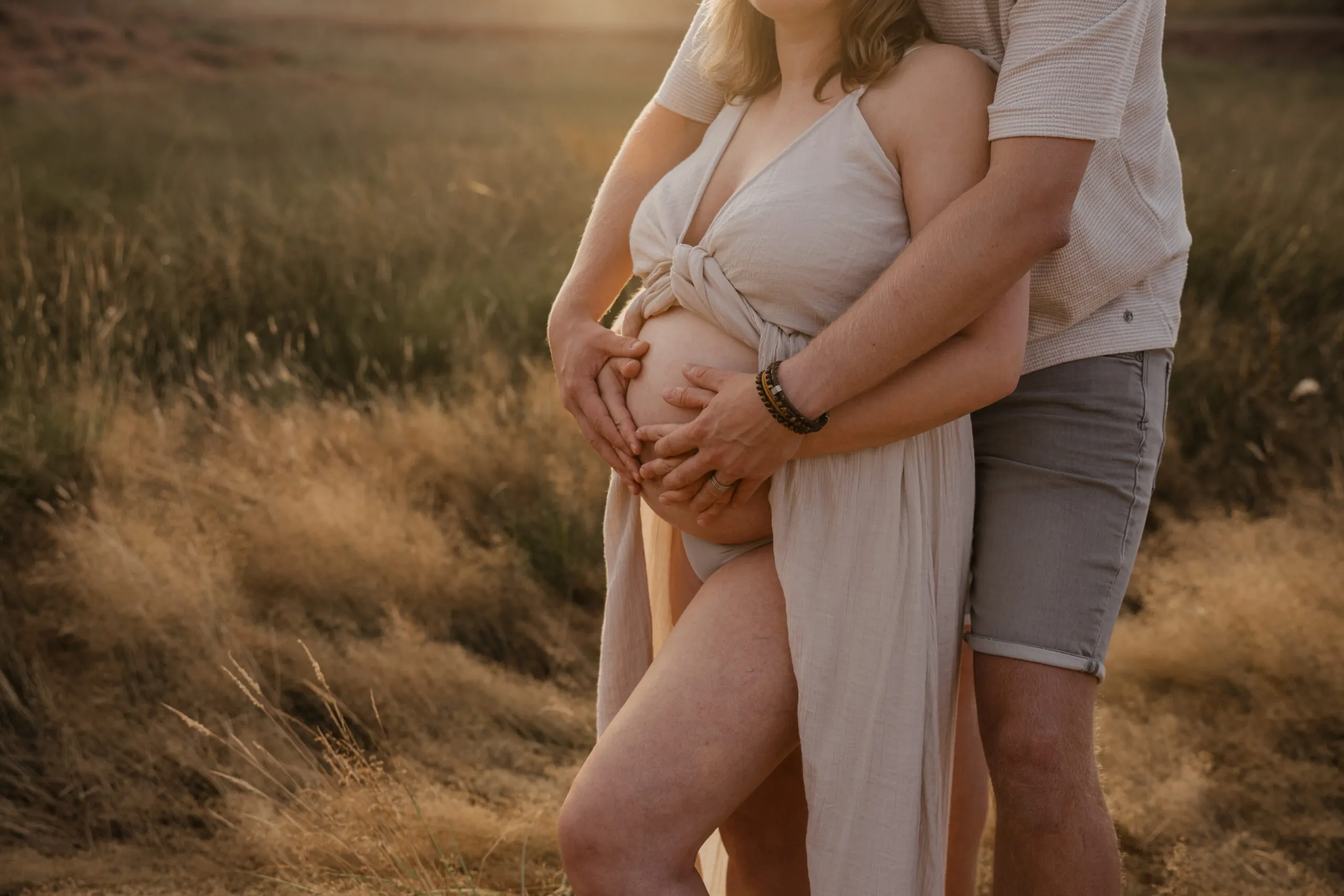 Handen van beide ouders om de zwangere buik in het hoge gras